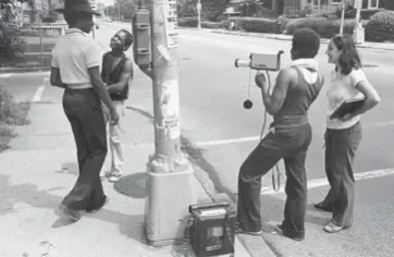 Old photo (likely 70s) of mixed race youth at a street corner. One is holding a video camera.