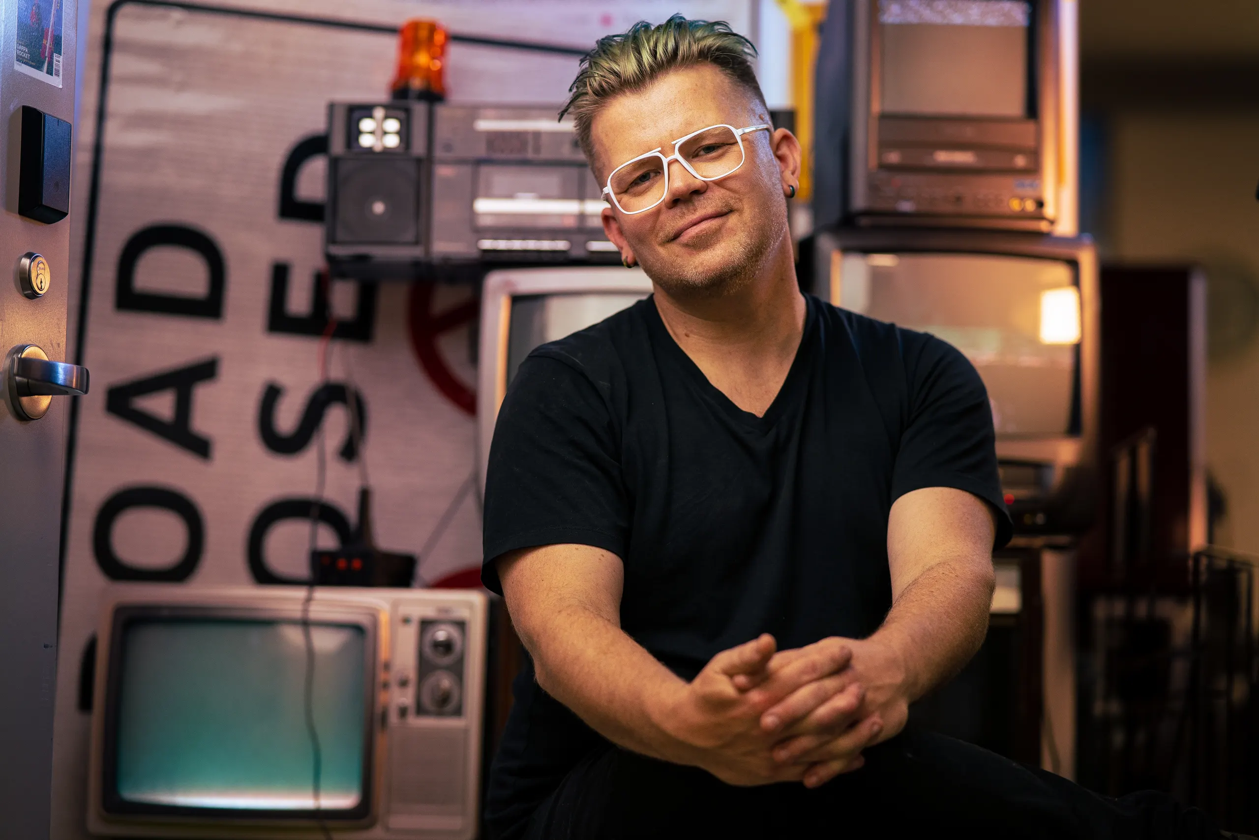 Portrait photo of Garnet Hertz with black t-shirt and half-smile, hands folded over the knees, surrounded by old televisions