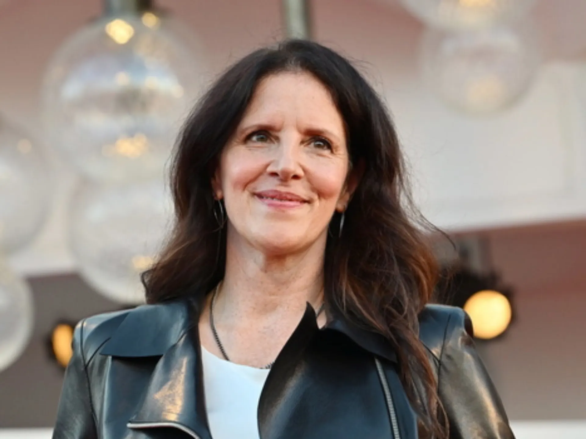 Photo of Laura Poitras, a woman with long dark hair wearing a black leather jacket over a white top, smiling and looking off to the side in front of blurred globe lights.