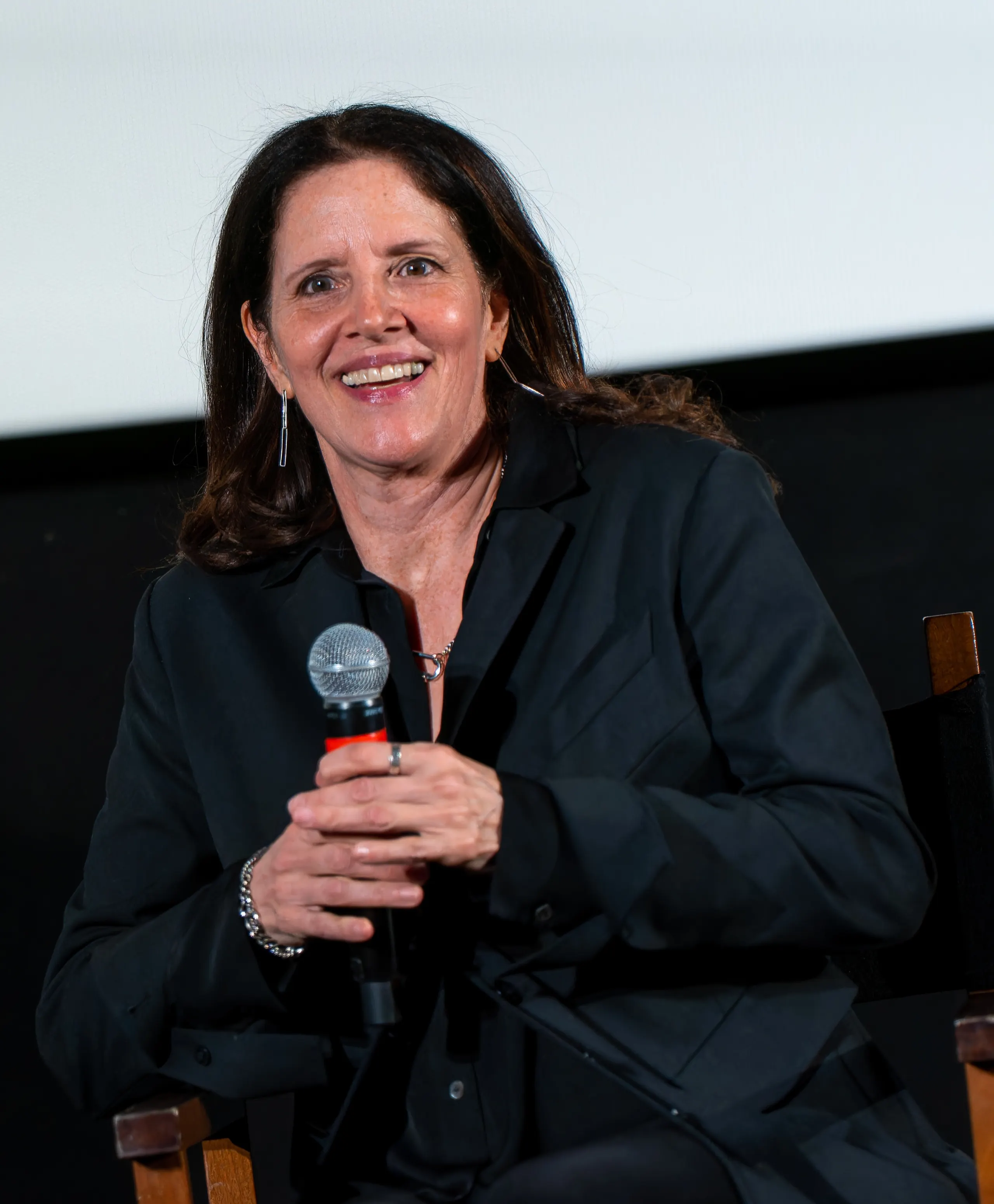 Photo of Laura Poitras, a woman with shoulder-length dark hair wearing a black jacket, smiling and holding a microphone while seated in front of a cinema screen.