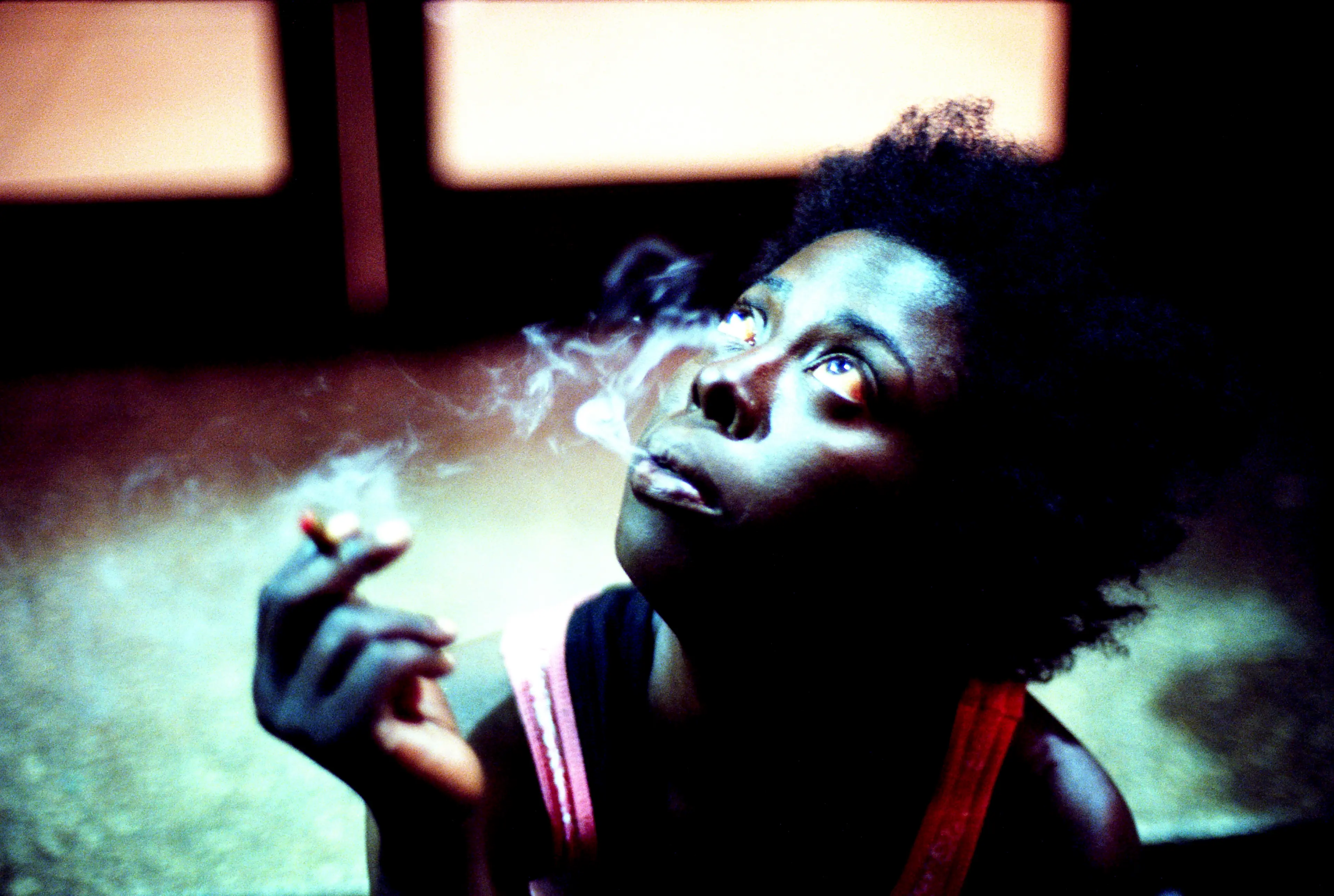 Photo of black woman with short hair in a dark surround, smoking, looking up towards light.
