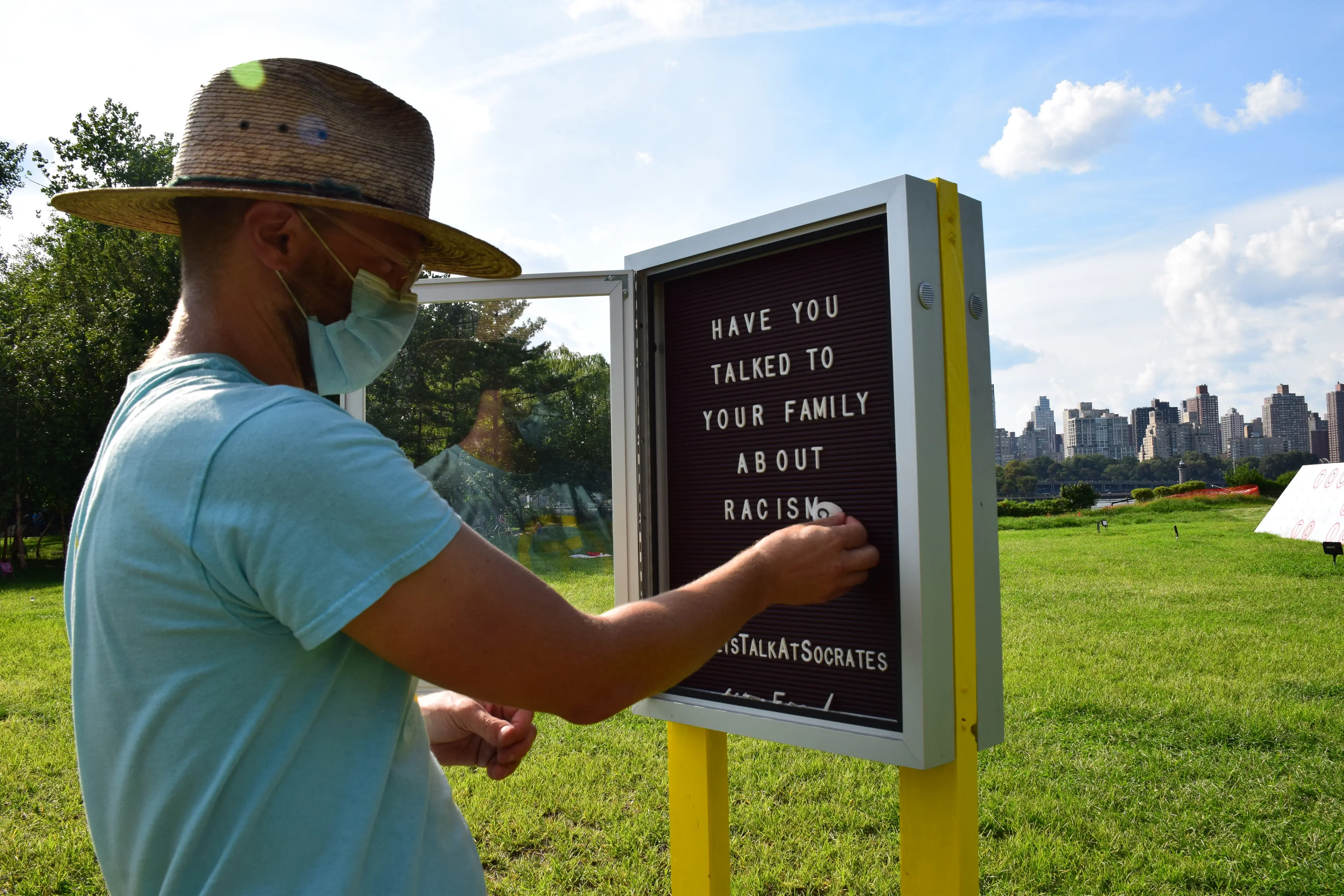 Photo of Doug wearing straw hat and covid mask arranging letters on a sign that says "Have you talked to your family about racism"