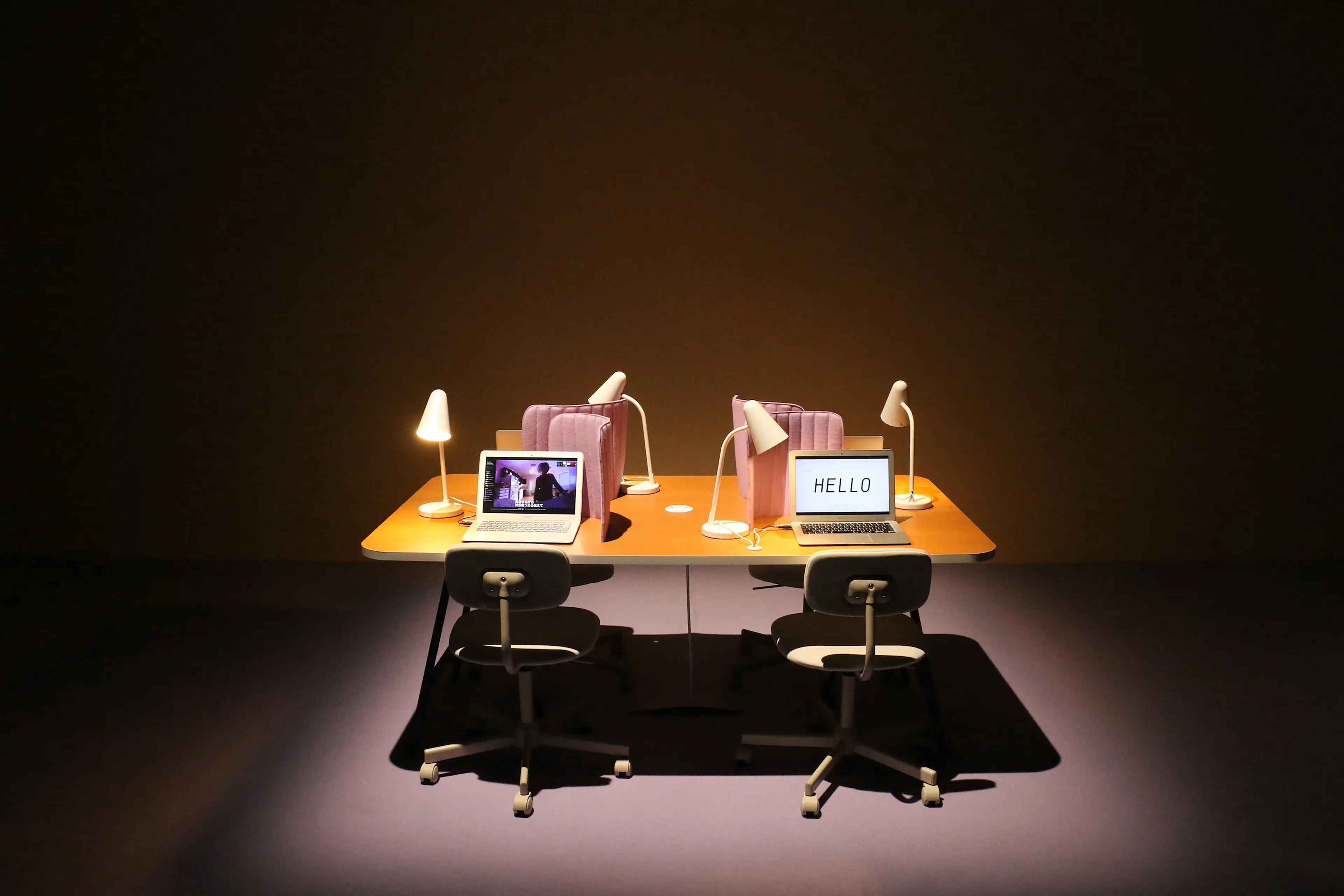 View of a desk and four empty chairs in a dark room lit from above.  Each chair has a laptop and a lamp on the desk.