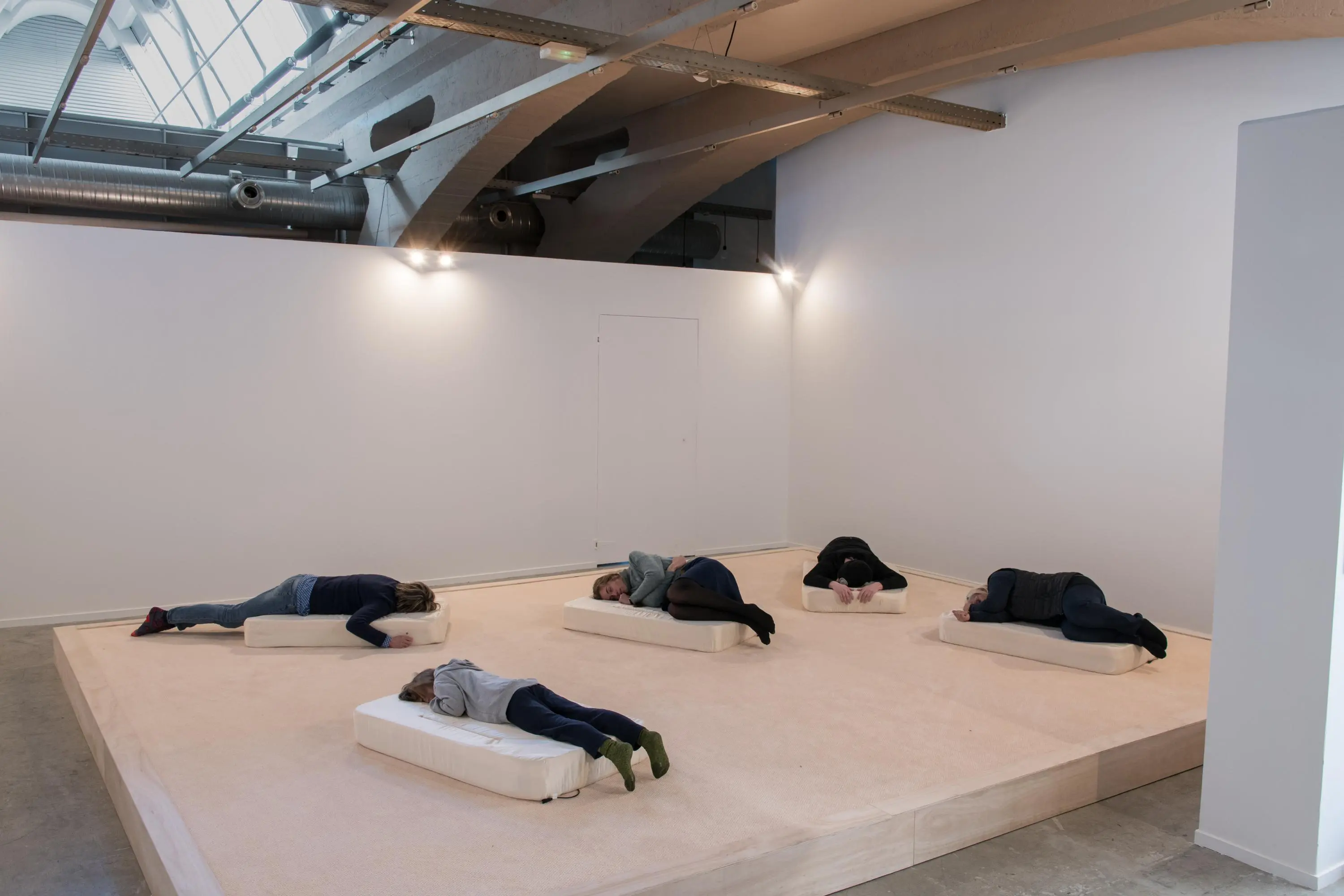 Five people in dark clothing laying on white matresses on top of a wooden platform in an otherwise empty gallery