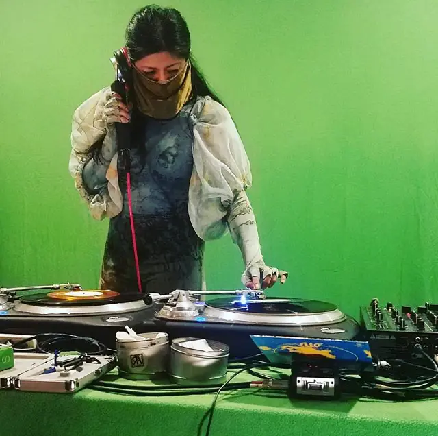 Photo of Maria with bright green background, wearing a white batik outfit covering her face and hands, standing and playing on two turntables.