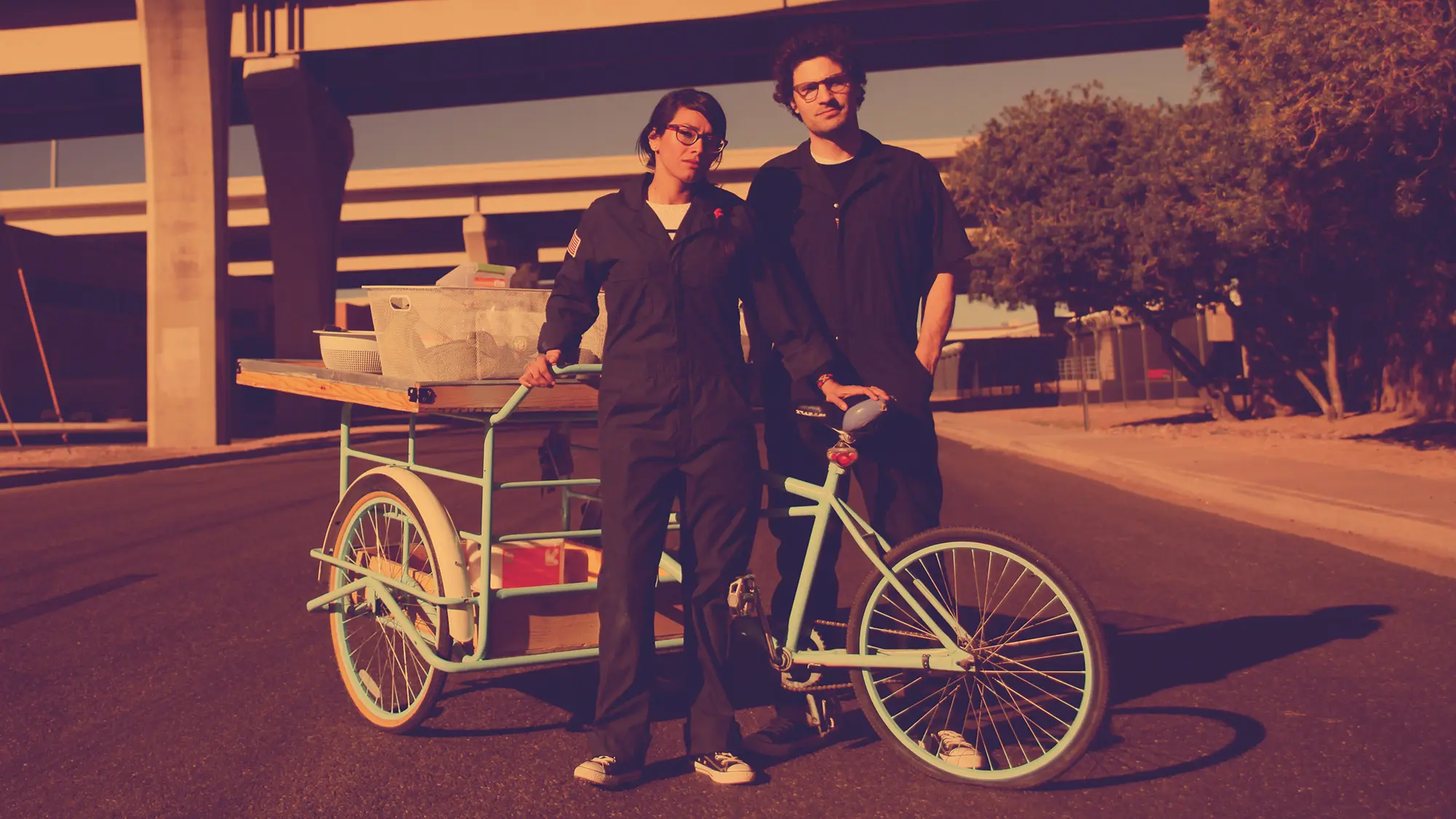 Photo portrait of Matt and April standing next to a three wheel light green cargo bike, both with dark blue overalls.