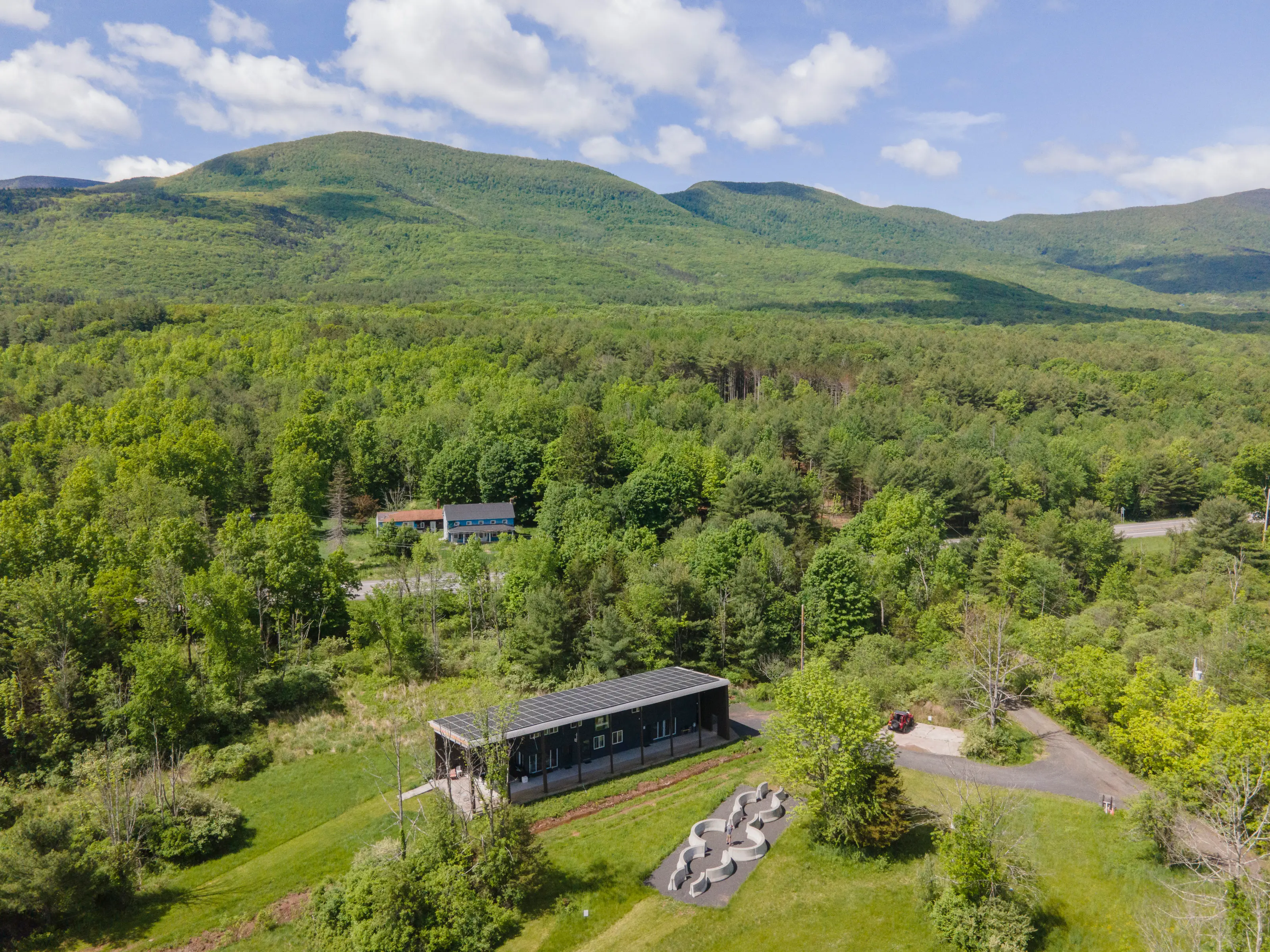 Ariel photo of Wave Farm showing a building with a solar roof, a large sculpture made of curved concrete barriers, and an appalachian landscape with a green forest and hills in the background.