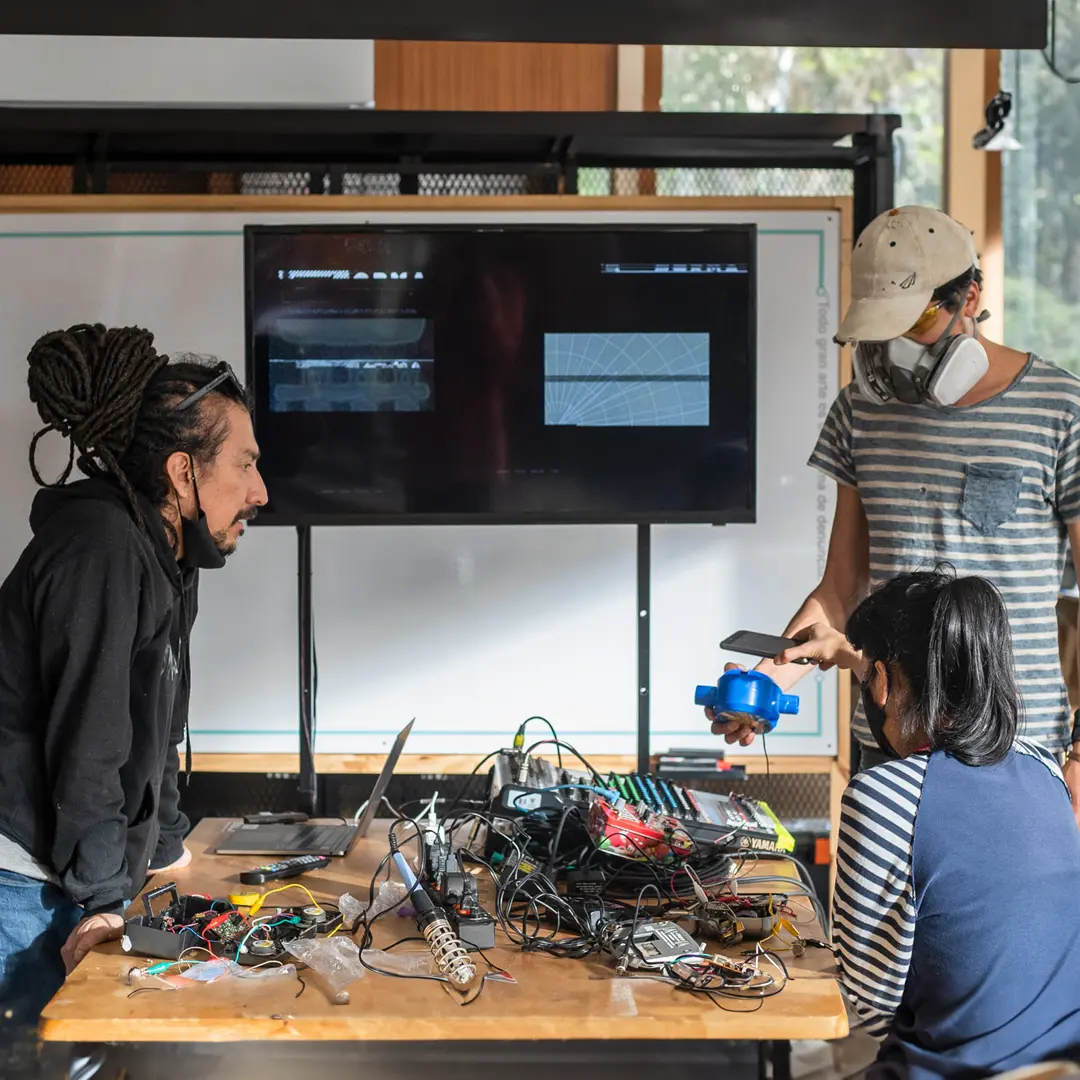 Three people sitting around a table full of electronics and cables. Man on the left has dreadlocks and is leaning with hands on desk.  The man int he background is wearing a large breathing mask and holding a blue container.  The women in the foreground has dark hair and is seated.