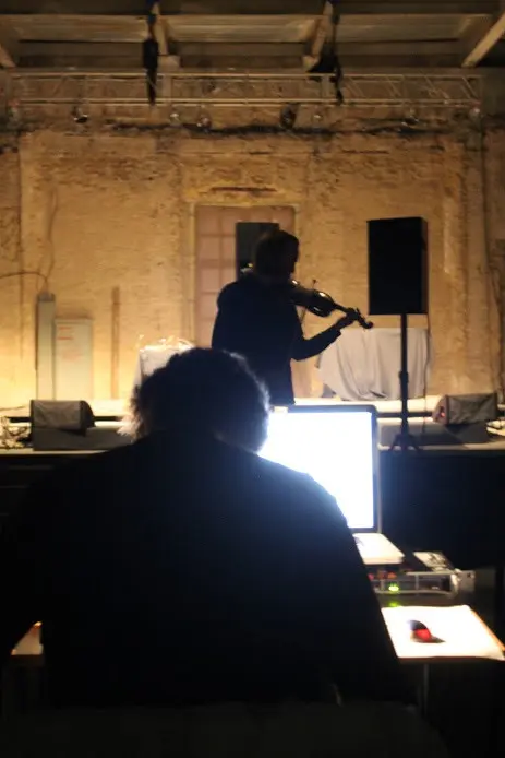 Photo of Schimana from behind, looking at a laptop, with violin player in background.