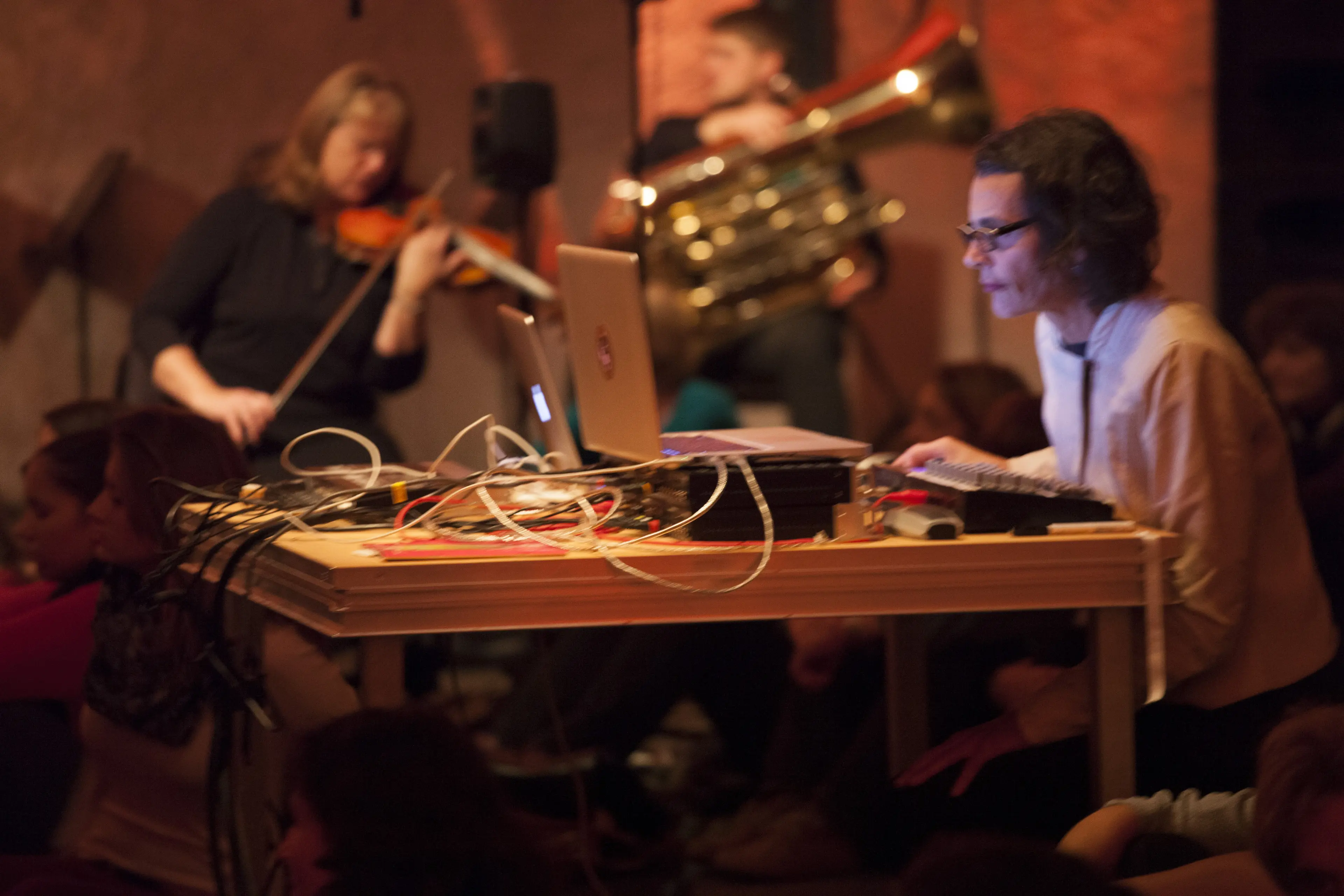Photo of Schimana looking to left at her laptop with violin and brass horn player in the background.