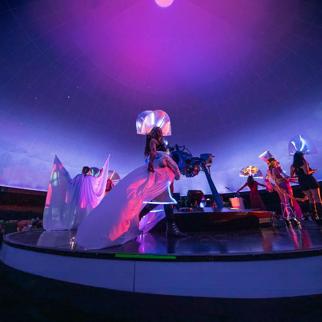 Performers in white futuristic costumes on stage in the middle of a planetarium.