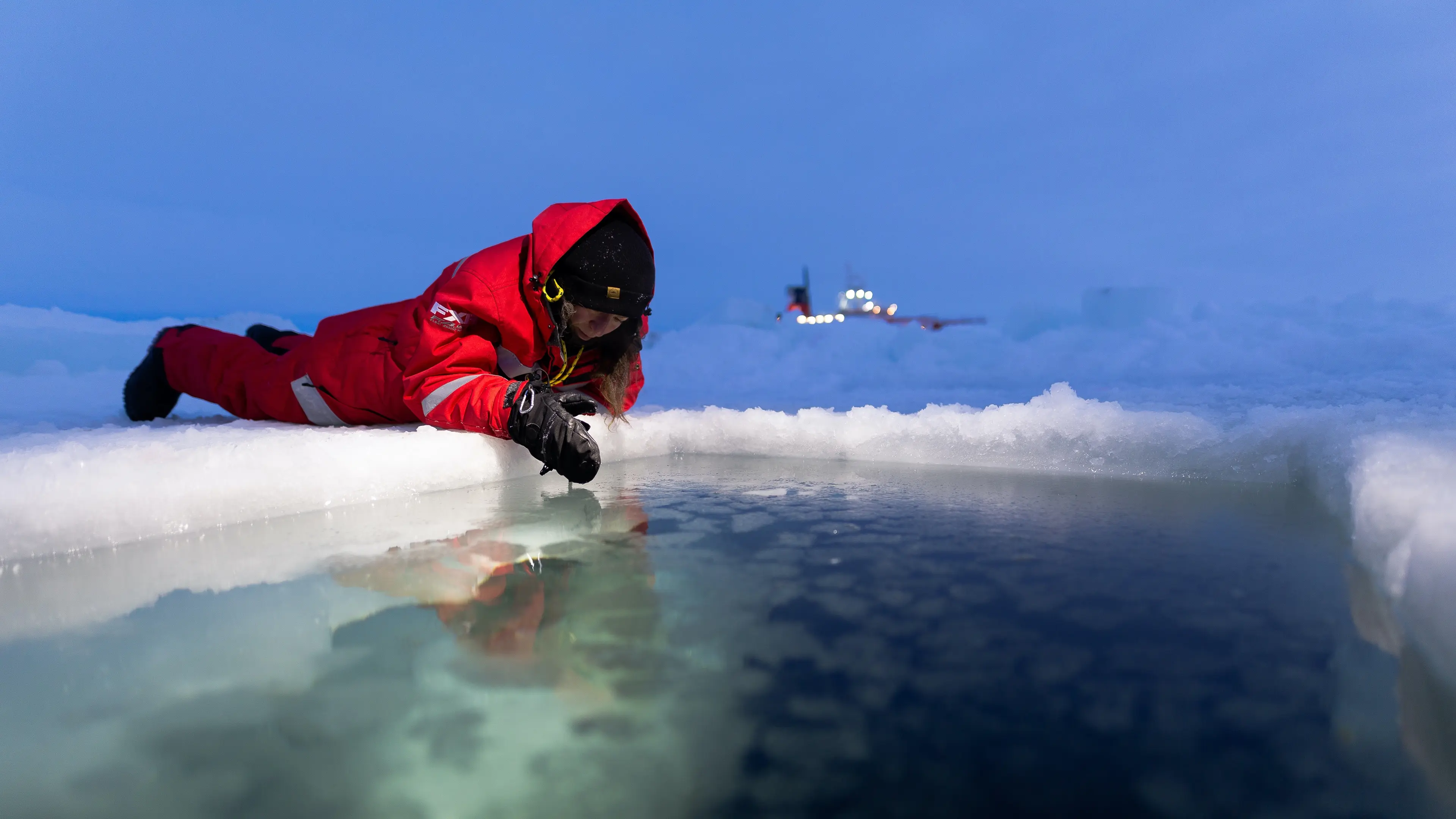 Photo of Amy in red winter suit laying face down on the ice with her face over a pool of water.