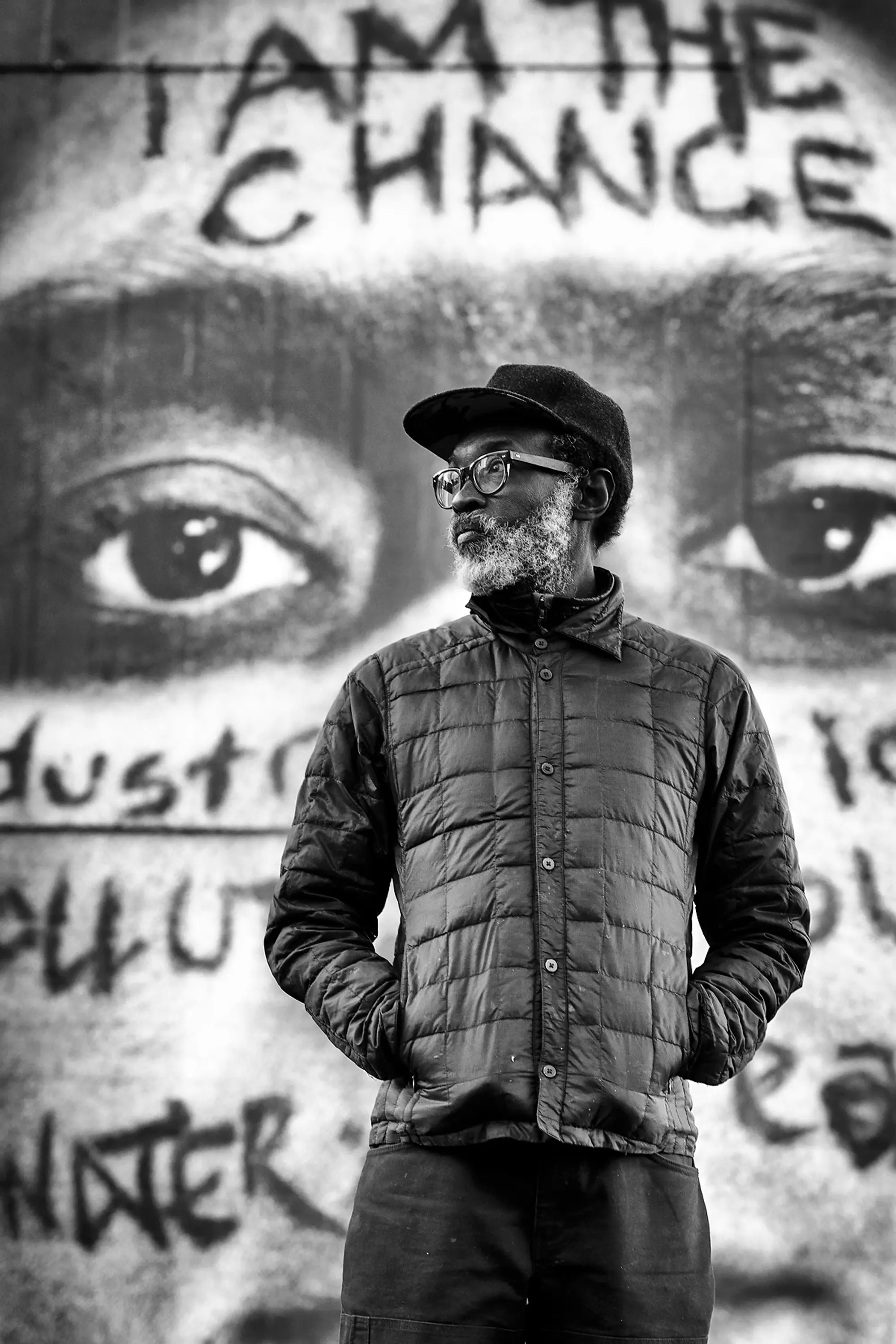 Photo portrait of Chip, a black man with white beard, glases, hat, and puffy jacket looking to the left. Behind him is a large photograph of a face.