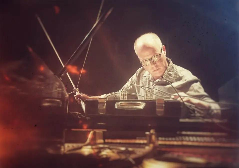 A white man with glasses and short hair sitting behind a stack of audio equipment and holding an antenna, lit from below and above.