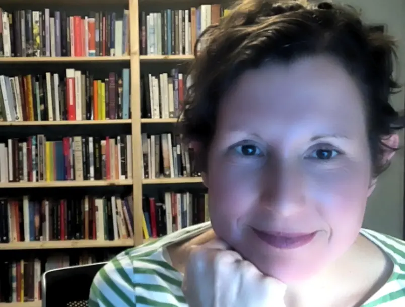 Photo portrait of Shannon, a white woman with dark hair and eyes, framed almost entirely on the face.  She is looking straight at the camera with a soft smile, resting her face on her right hand under her chin. She is wearing a green and white striped shirt.  In the background, we see a full bookshelf.