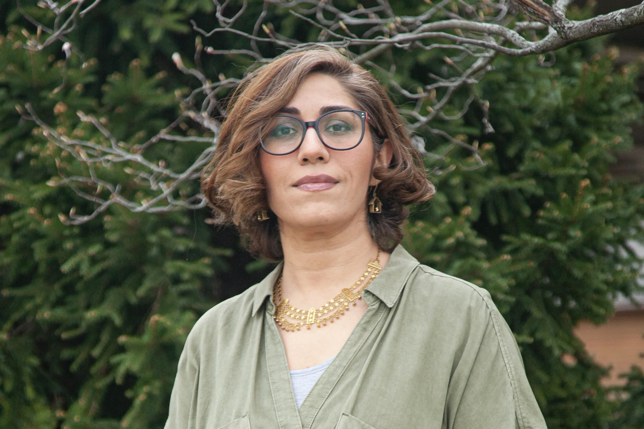 Photo portrait of Maryam outside with brown hair, large round glasses with a earth green blouse.