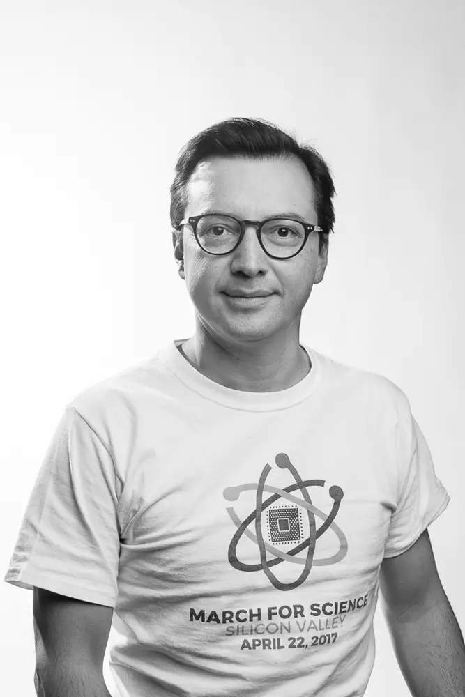 A dark-skin man with black hair and black glasses looking directly at the viewer and wearing a white tshirt that says "Marching for Science"