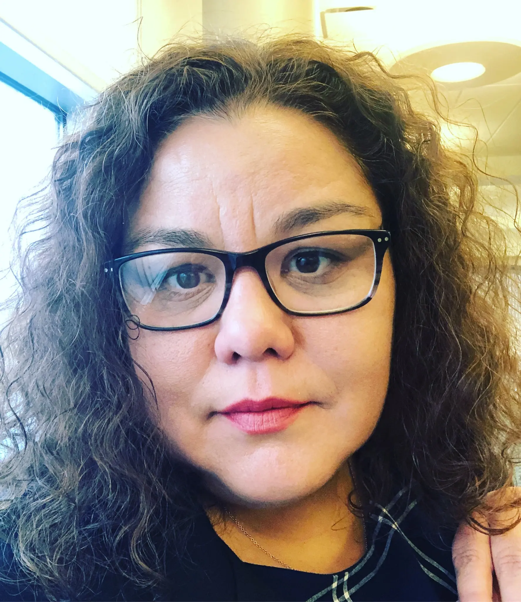 white woman with dark black hair and a reddish stripe on the left side, wearing glasses and liptstick and necklace with a circle-over-cross pendant.