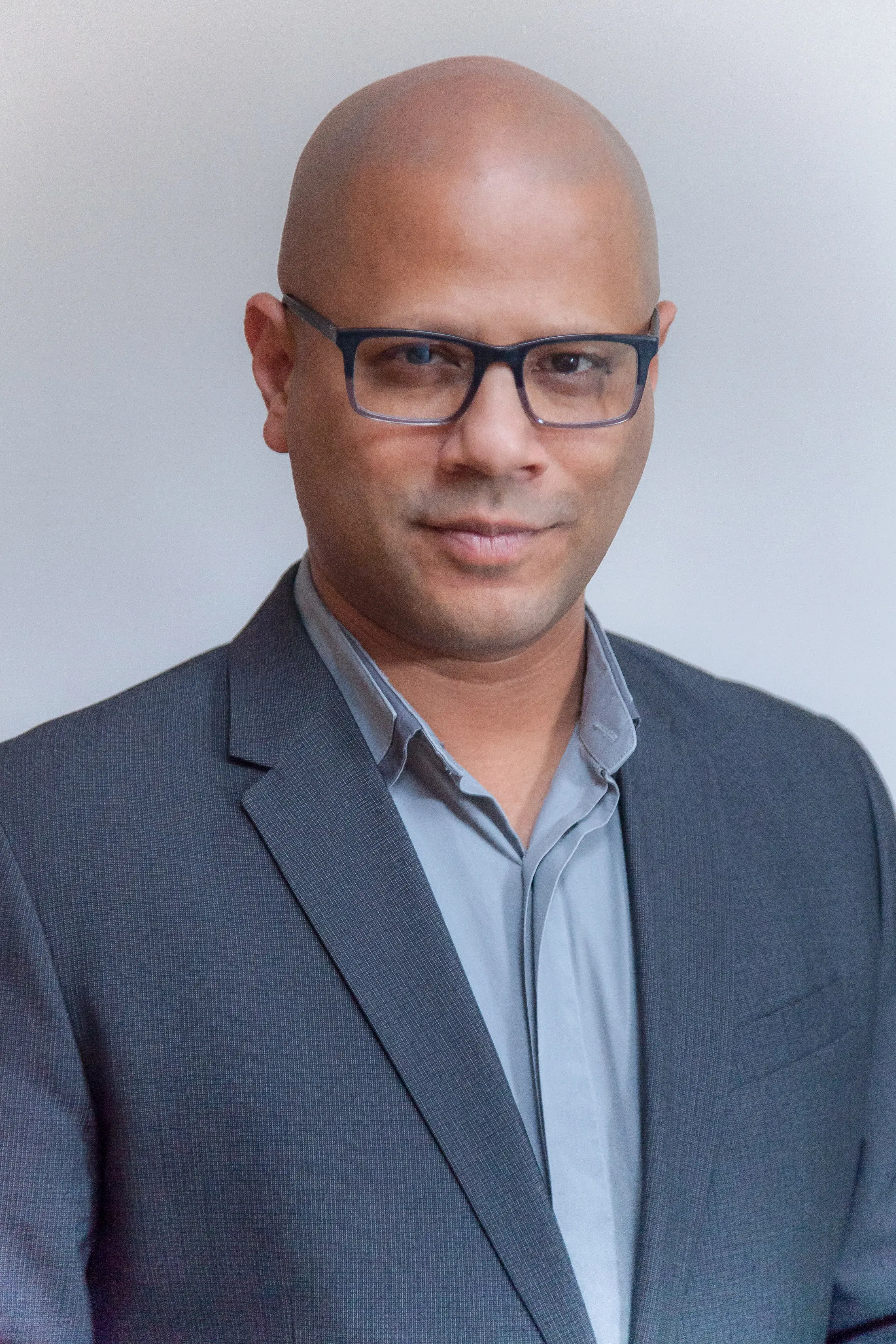 Color portrait photo of Hasan in light grey shirt with grey blazer, a shaved head and dark glass with a slight smile looking directly at the camera.