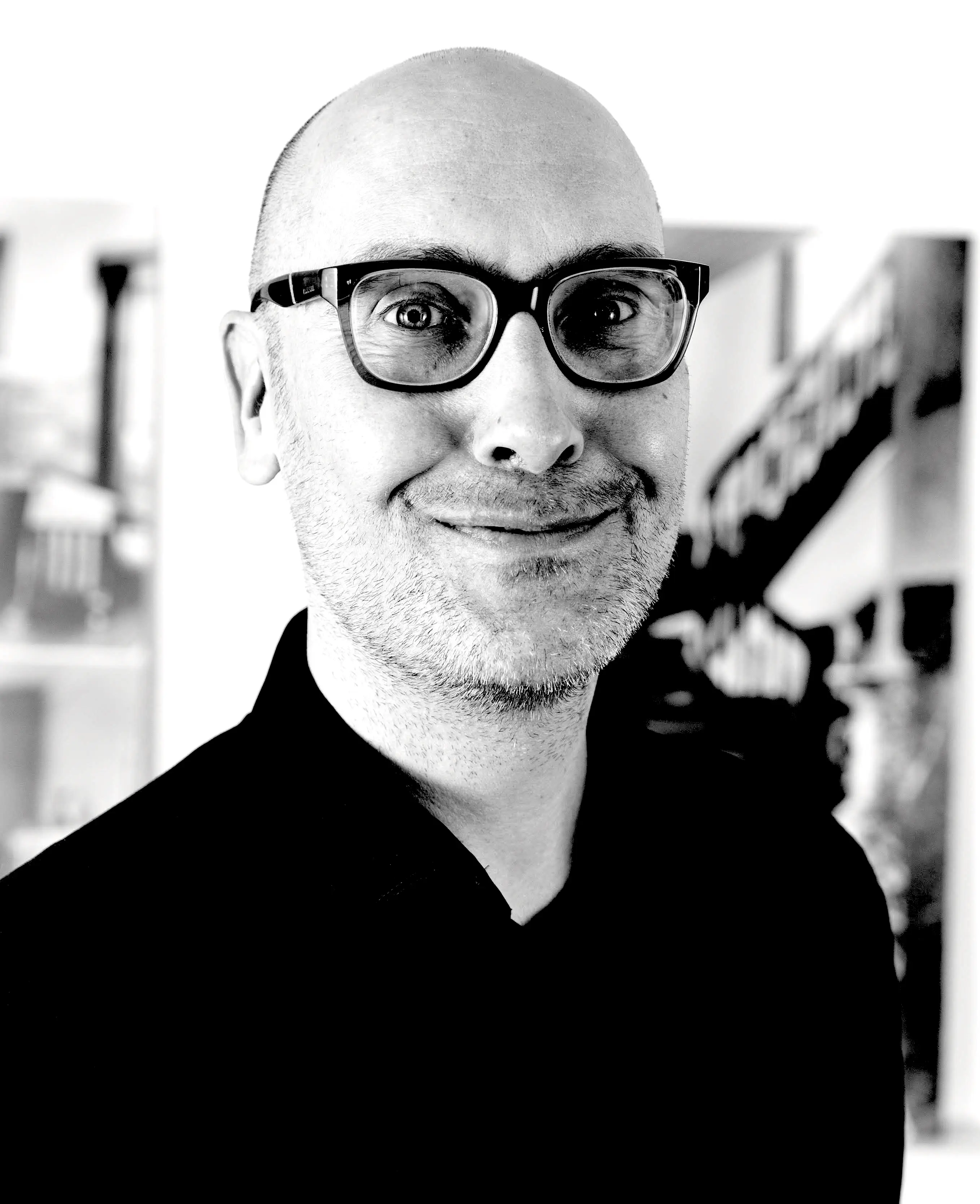 Black and white photo of Jussi Parikka in black shirt, dark thick glasses with a shaved head and a smile.
