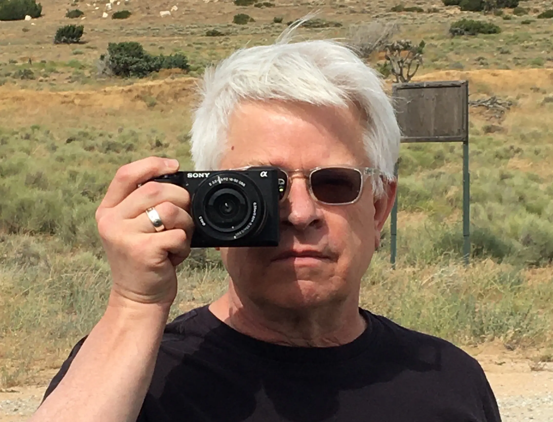 A white man with white hair and black t-shirt, wearing sunglasses and holding a camera over his left eye pointed at the viewer.