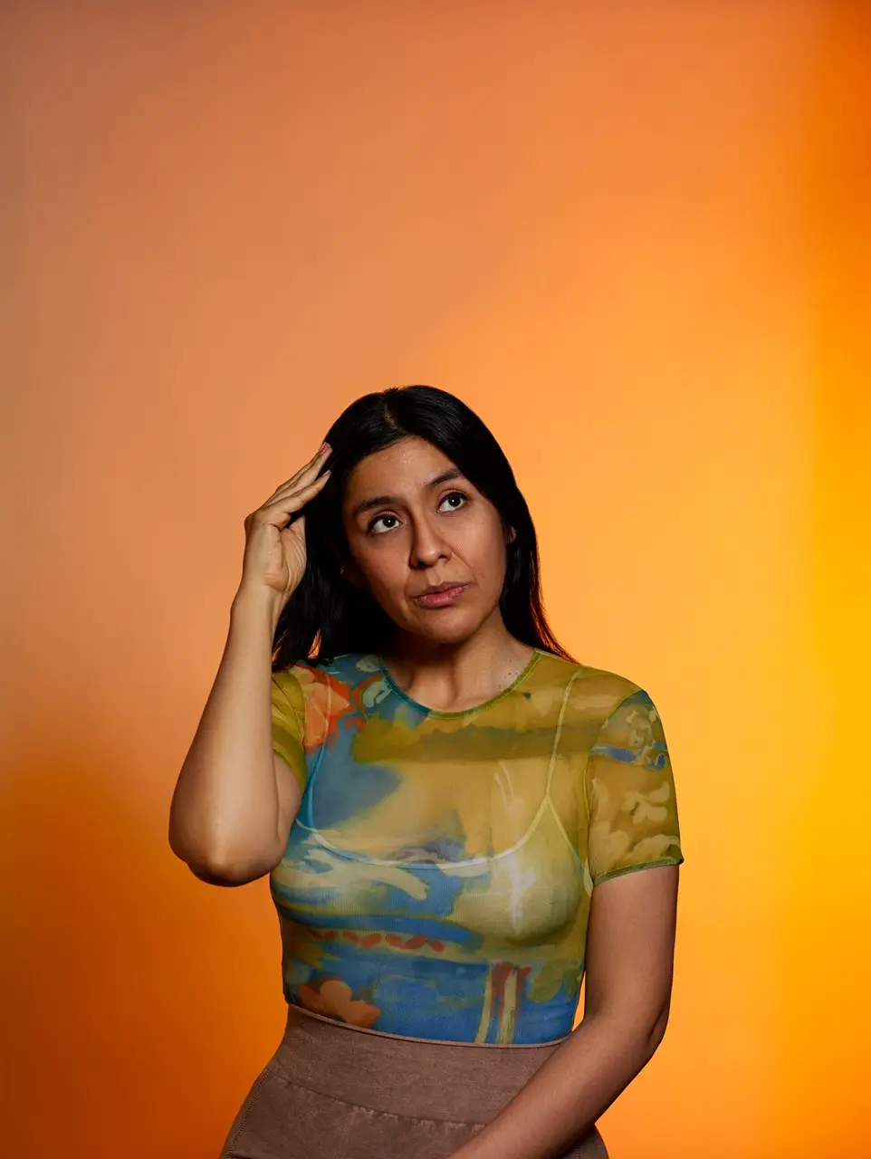 Portrait photo of Maria with long black hair and orange background, wearing partially see-through printed shirt, looking upwards with mischeavous smile.