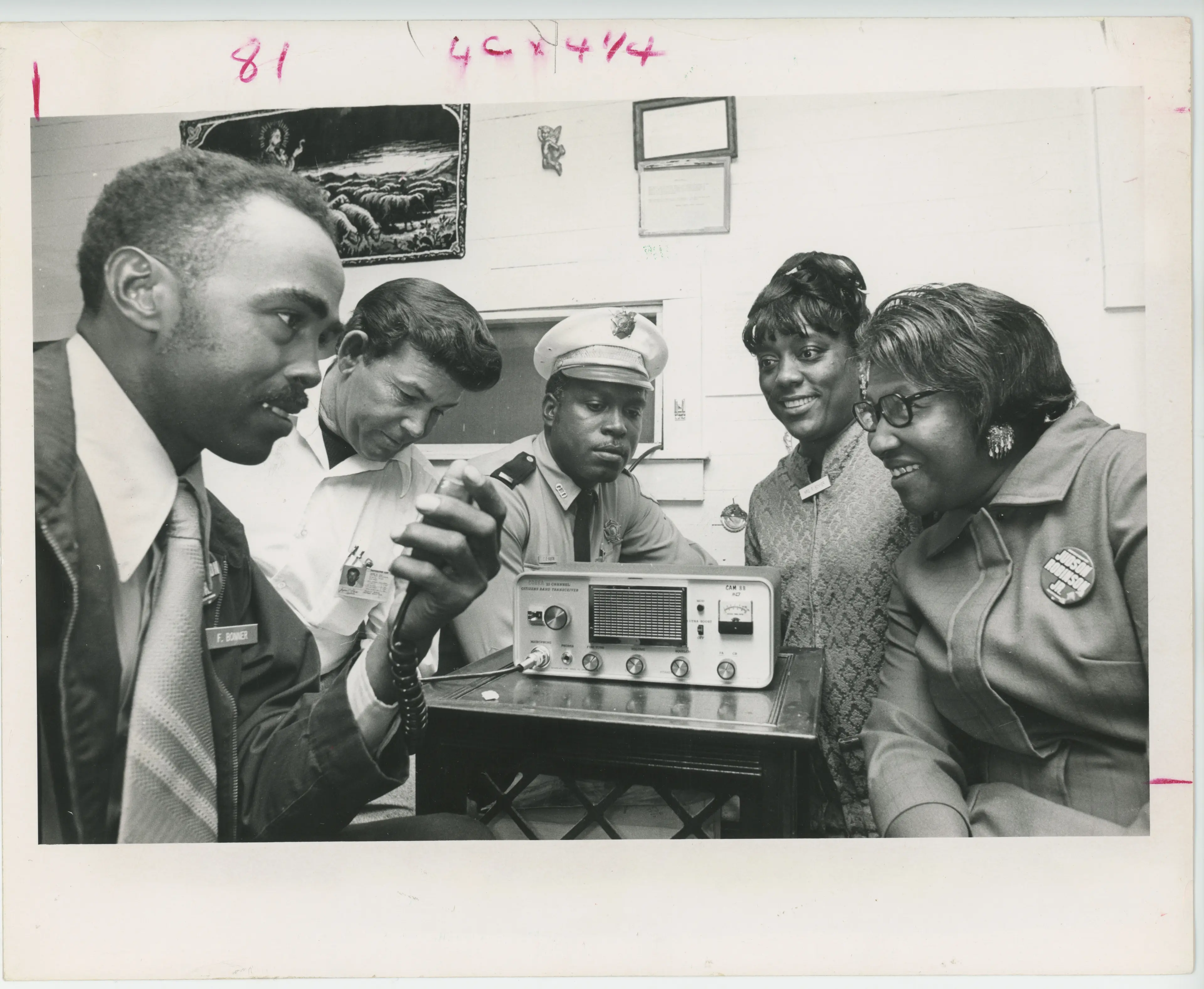 Black and white photo of three black men and three black women sitting around a CB radio with the man in foreground speaking into the microphone