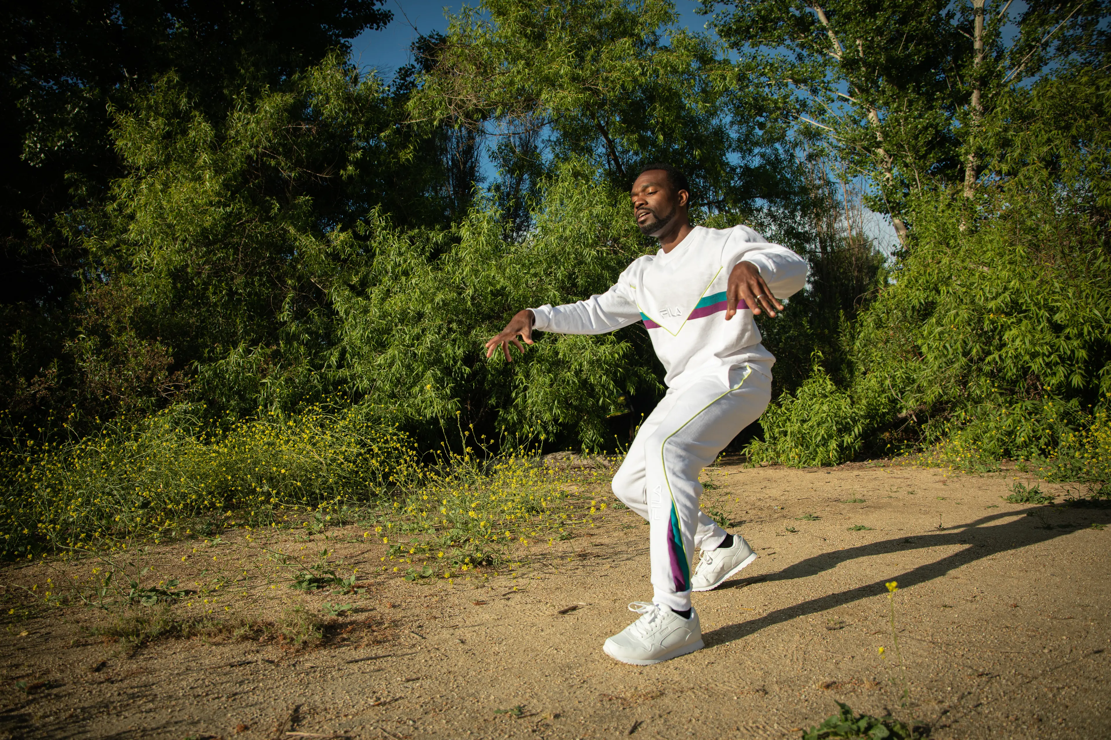 Photo of black man (André) in white attire dancing on a dirt trail with deciduous trees in the background.
