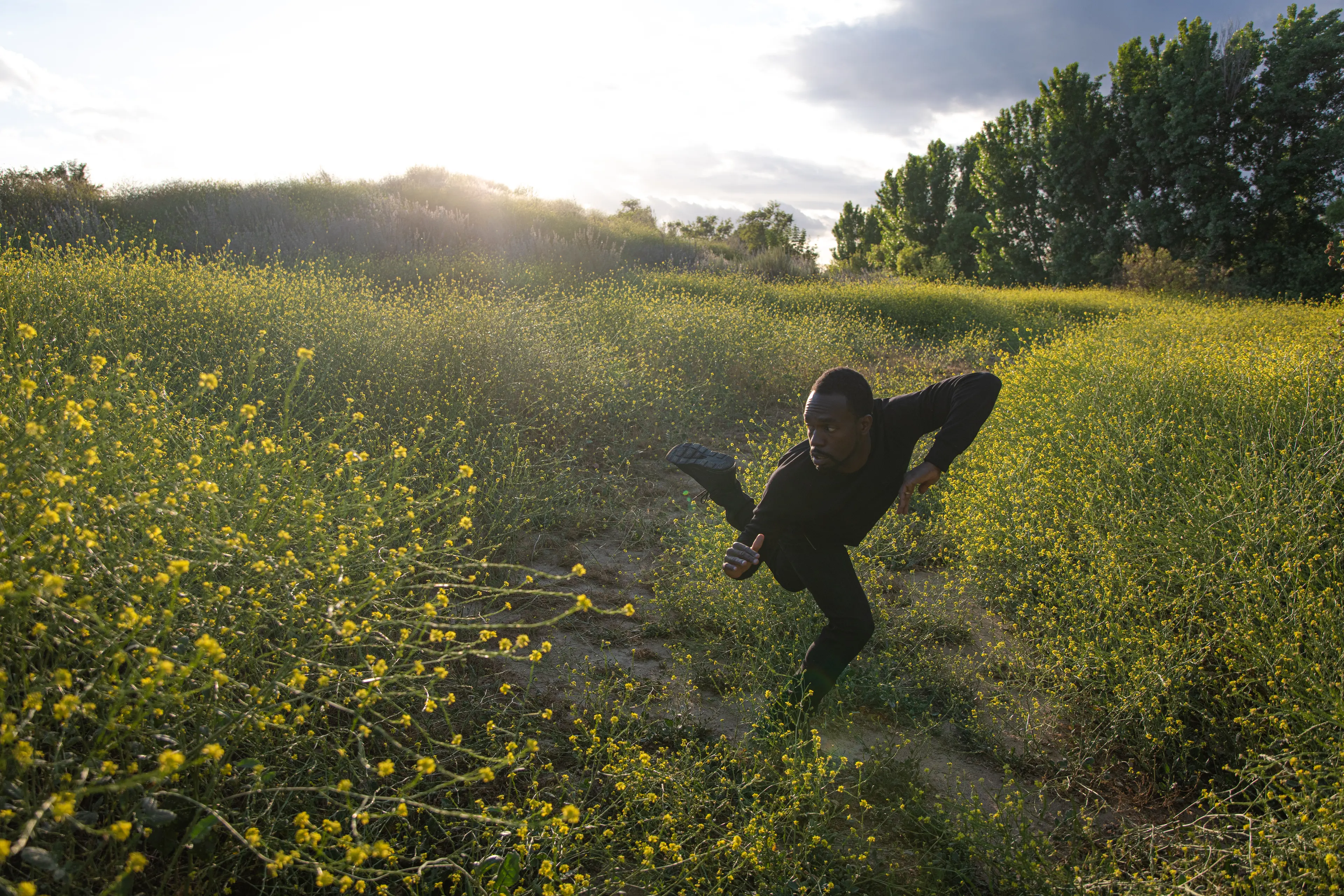 Photo of black man (André) in black attire dancing in a field of yellow flowers with the sun behind in the horizon.